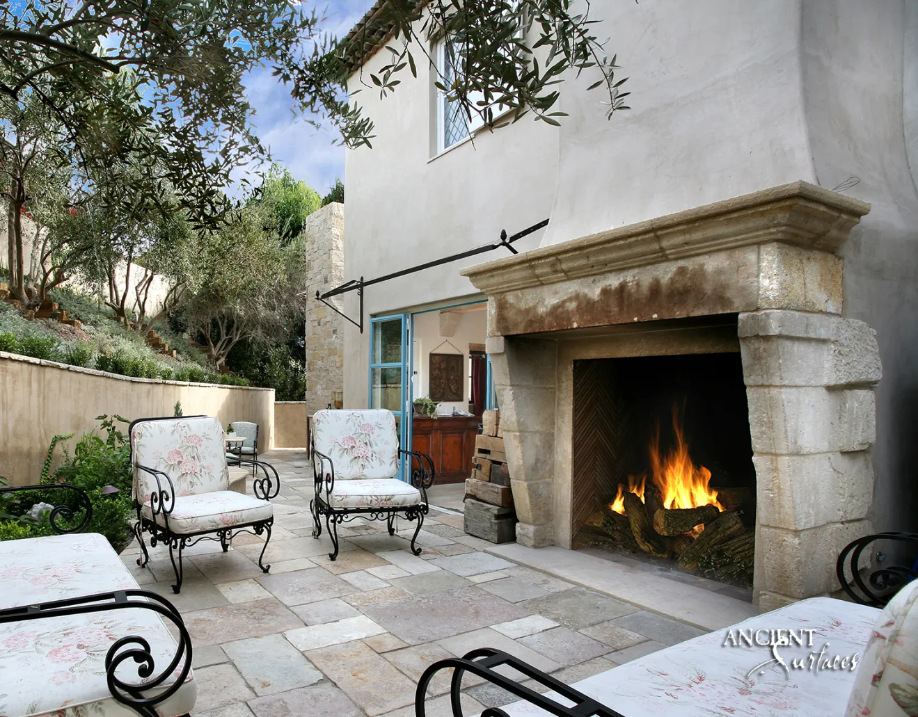Monumental reclaimed limestone fireplace mantel from Ancient Surfaces with a deep uncarved lintel on battered block jambs, installed on a Mediterranean terrace with a lit herringbone brick firebox