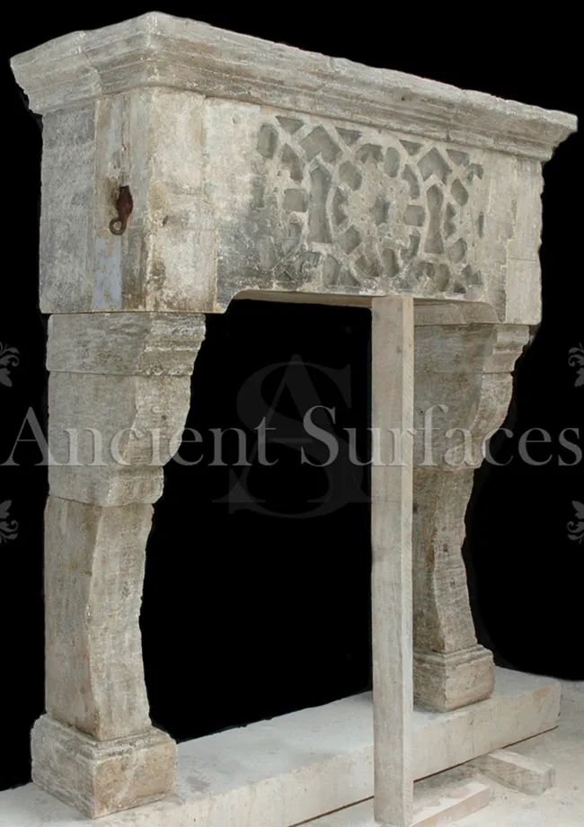 Reclaimed antique limestone fireplace mantel from Ancient Surfaces with an openwork pierced strapwork frieze and deep scrolled corbel jambs, photographed against a black studio background