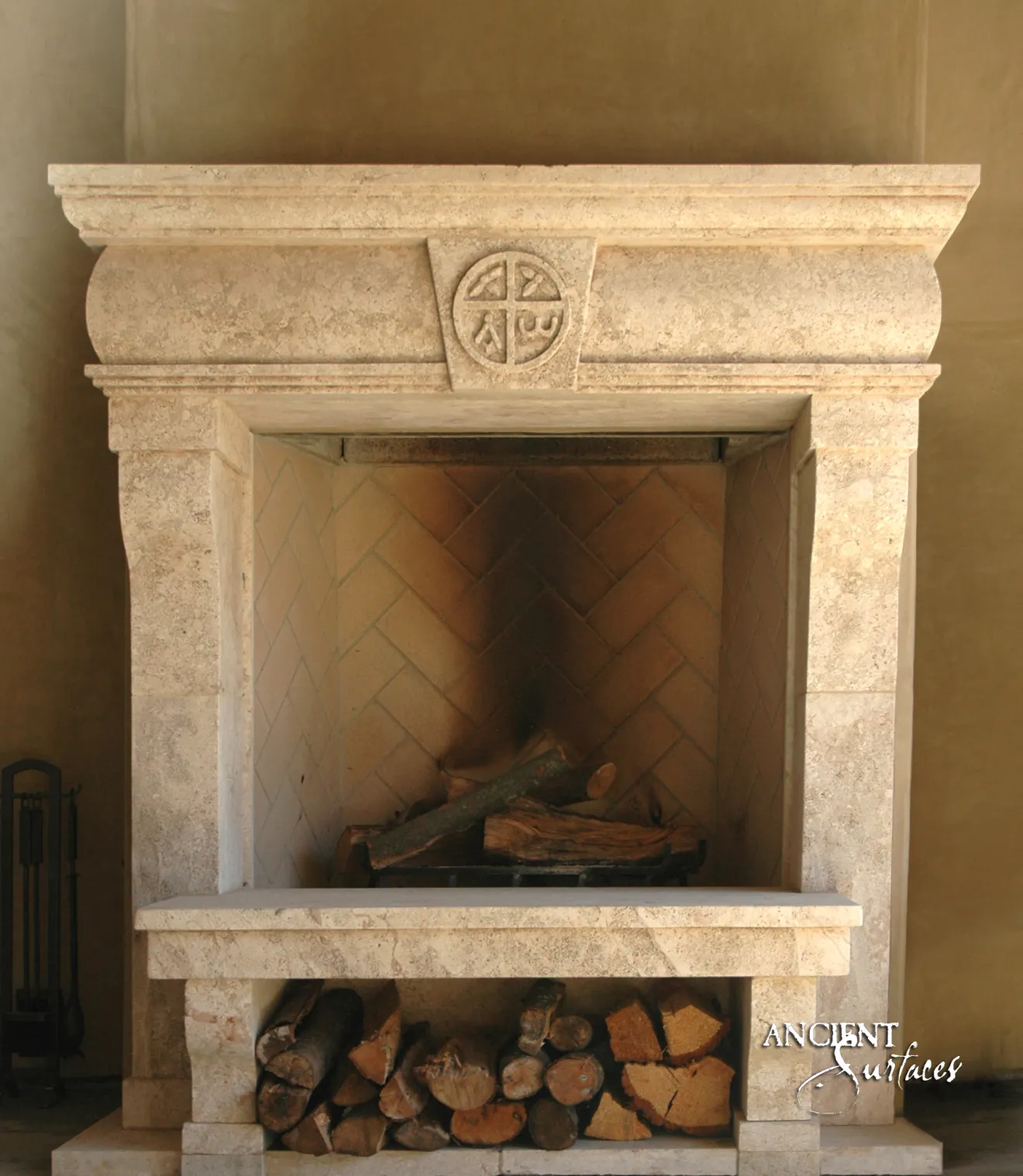 Reclaimed antique limestone fireplace mantel from Ancient Surfaces with a cushioned frieze and carved wheel roundel above a herringbone firebox and an open log store, against an ochre plaster wall