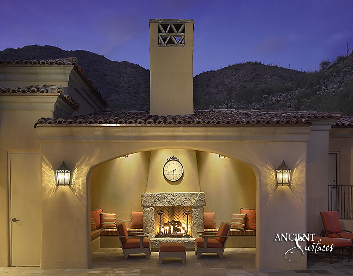 Rough hewn reclaimed limestone fireplace from Ancient Surfaces with a single undressed lintel on stone piers, burning at dusk in a tiled roof desert loggia below the mountains