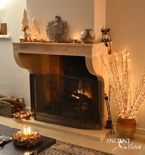 Reclaimed antique French limestone fireplace mantel from Ancient Surfaces with carved rosettes and a ribbon frieze above long scrolled jambs, behind an iron fire screen in a plain contemporary room