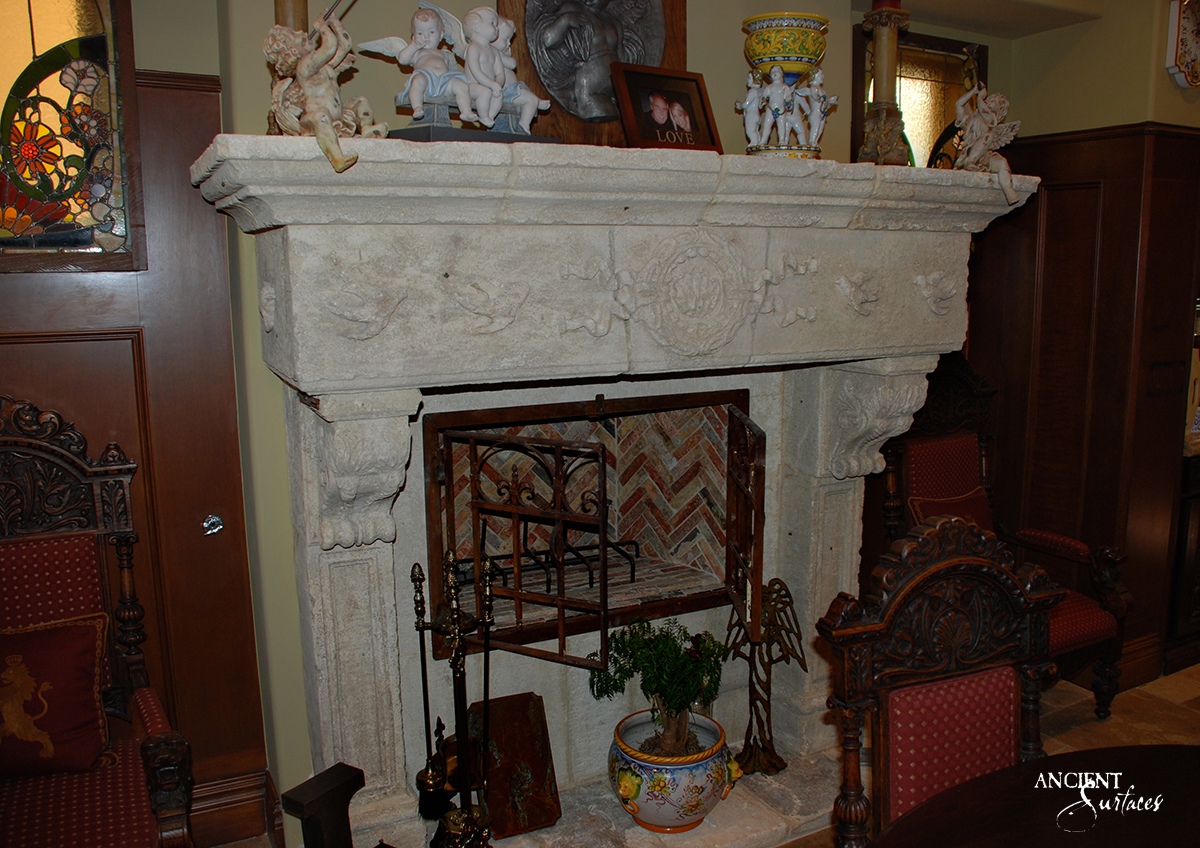 Reclaimed antique limestone fireplace mantel from Ancient Surfaces carved with facing doves and swags around a laurel wreath, in a wood panelled room with stained glass and a red brick herringbone firebox