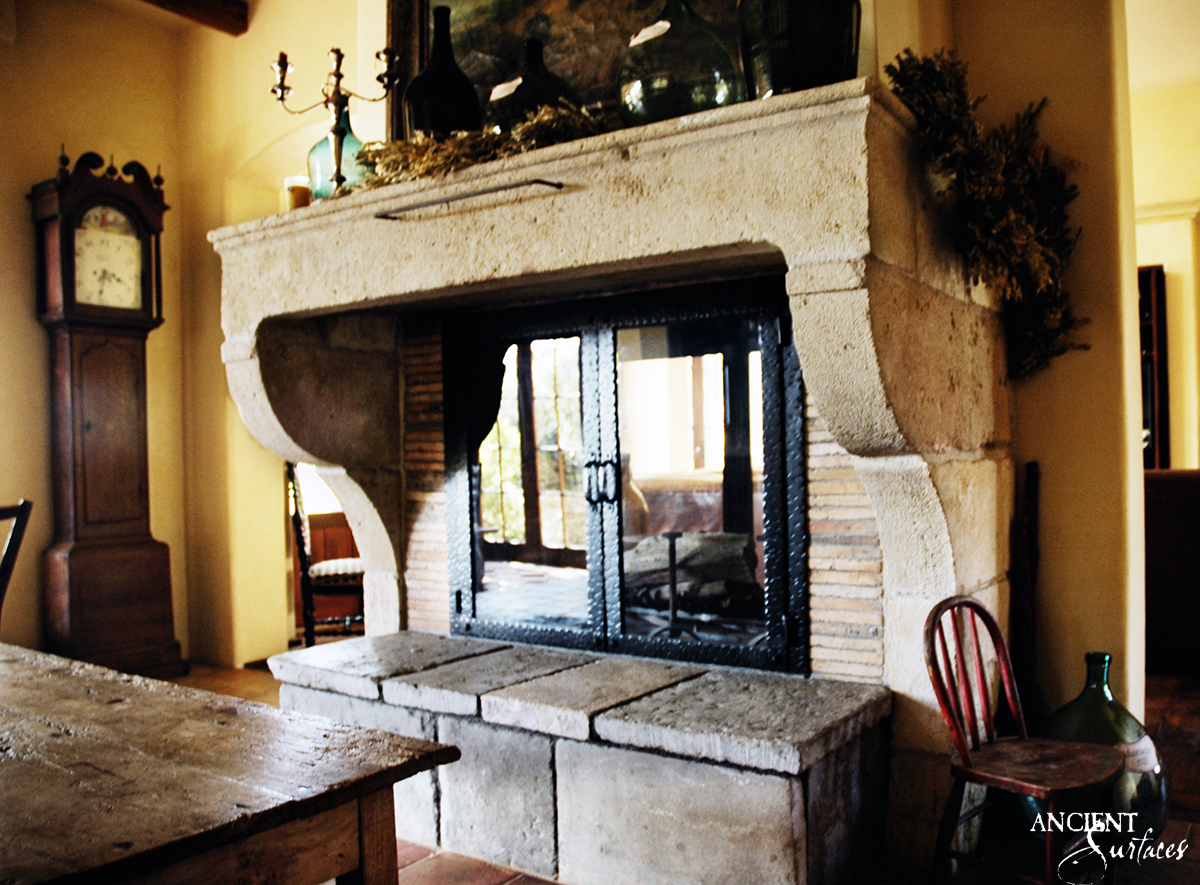 Reclaimed antique limestone fireplace mantel from Ancient Surfaces with deep scrolled consoles and rope twist iron doors over a double sided firebox, in a warm ochre kitchen beside a longcase clock