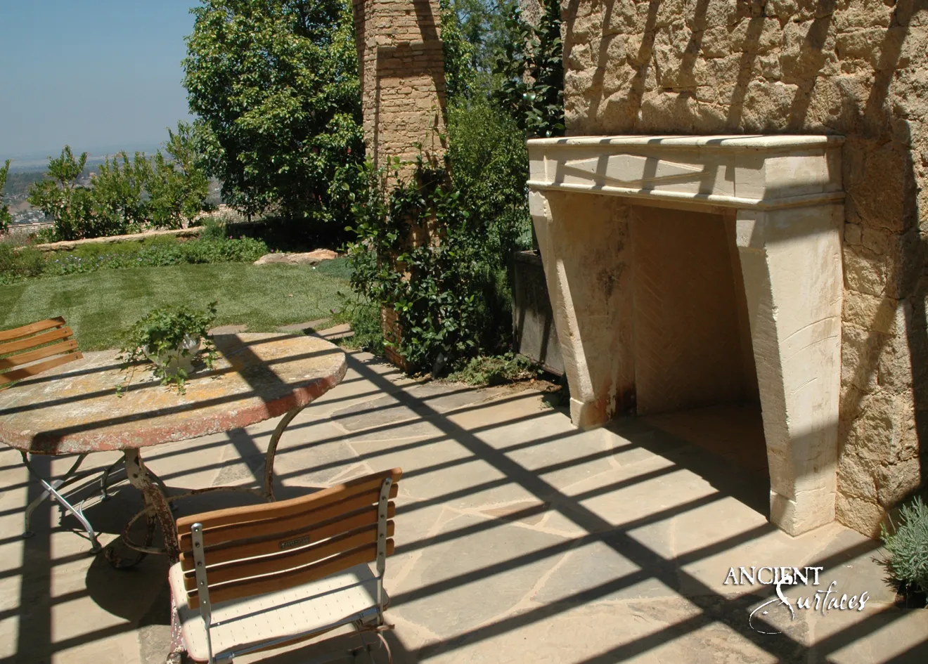 Sun bleached reclaimed limestone fireplace mantel from Ancient Surfaces built into a rubble stone garden wall, on a hilltop terrace striped with pergola shadows
