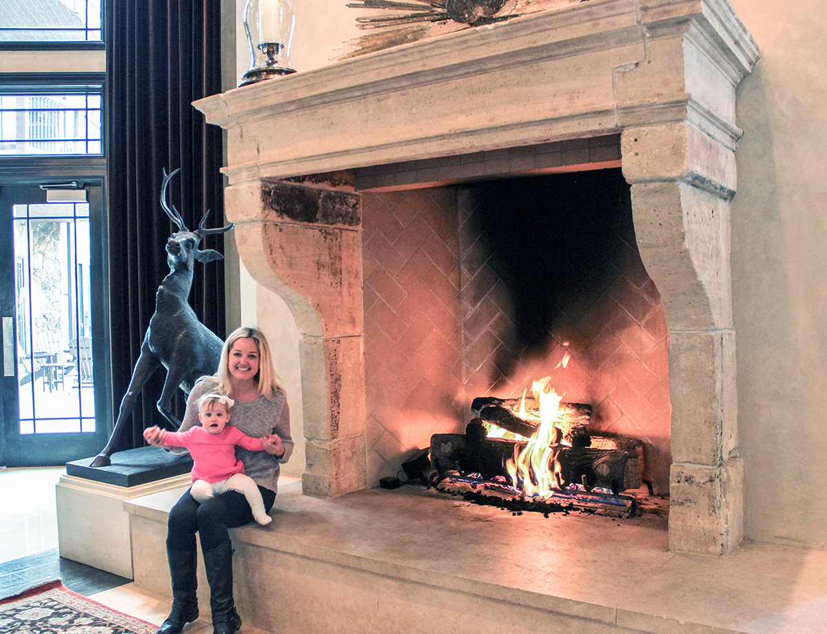 Oversized reclaimed limestone fireplace mantel from Ancient Surfaces with deep scrolled console jambs and a broad raised hearth, burning in a double height great room