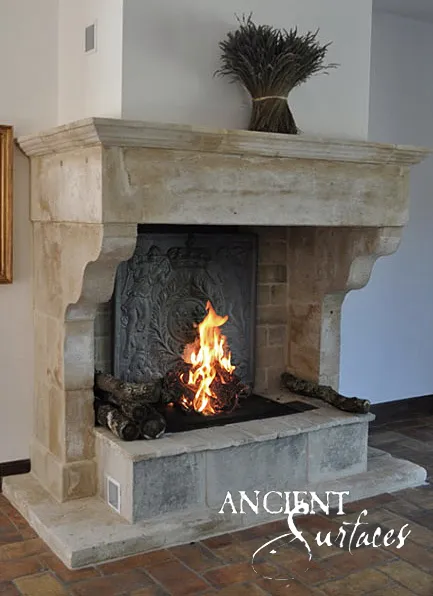 Freestanding reclaimed limestone fireplace from Ancient Surfaces with scrolled corbels on a stepped stone hearth platform, burning against a white wall over a terracotta tiled floor