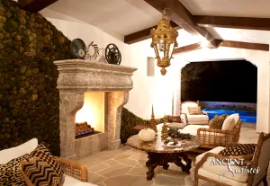 Compact grey reclaimed limestone fireplace mantel from Ancient Surfaces with scrolled corbel jambs, lit after dark against a moss covered wall in a poolside pavilion