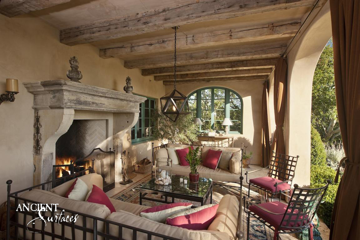 Grey white reclaimed limestone fireplace mantel from Ancient Surfaces with a stepped cornice and deep console jambs, alight in a covered outdoor room under massive reclaimed timber beams